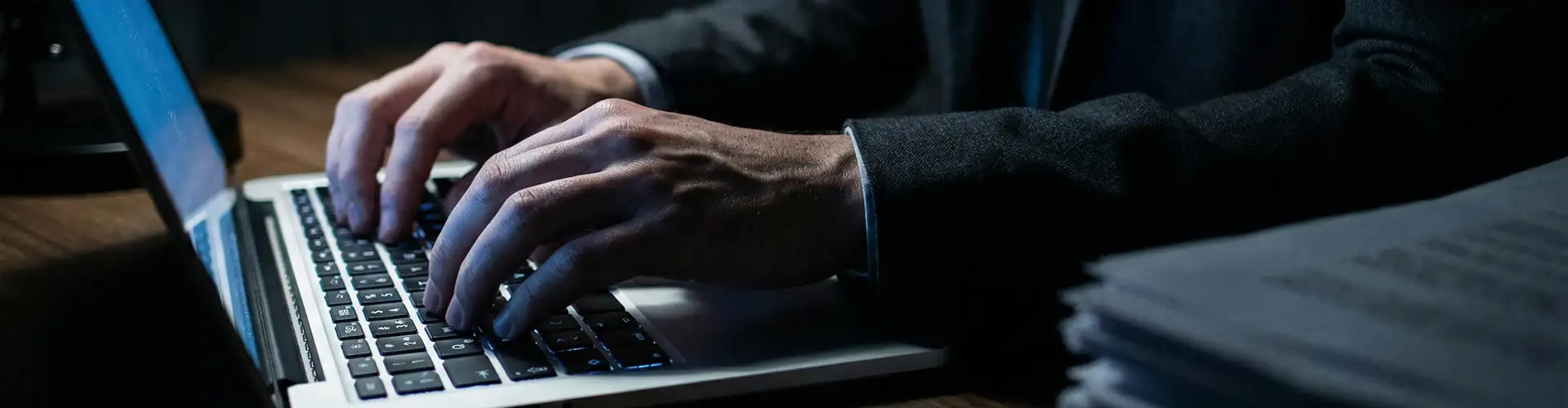 Dettaglio delle mani di un professionista in abito scuro che digita su un laptop per l'invio della dichiarazione di successione on line, con una pila di documenti cartacei a lato che simboleggia il portale agenzia delle entrate successione on line